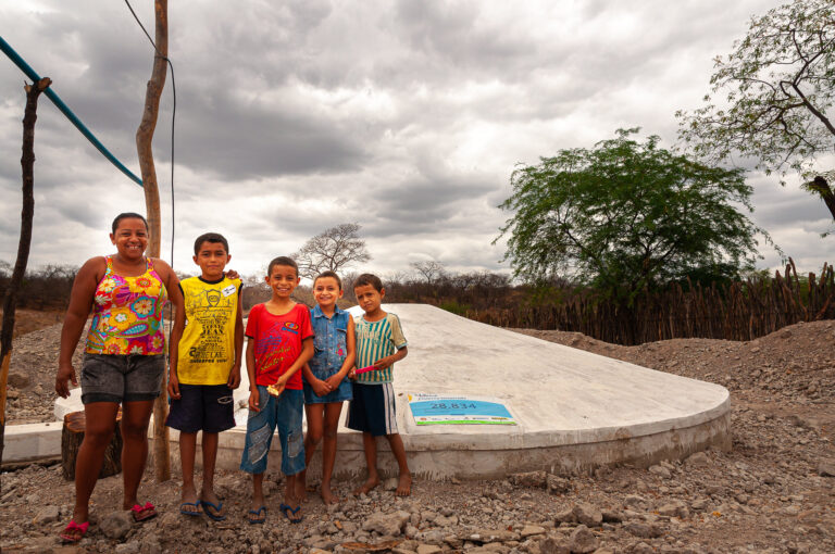 Mulher com quatro crianças sorridentes pousam para foto na frente de sua cisterna de segunda água.