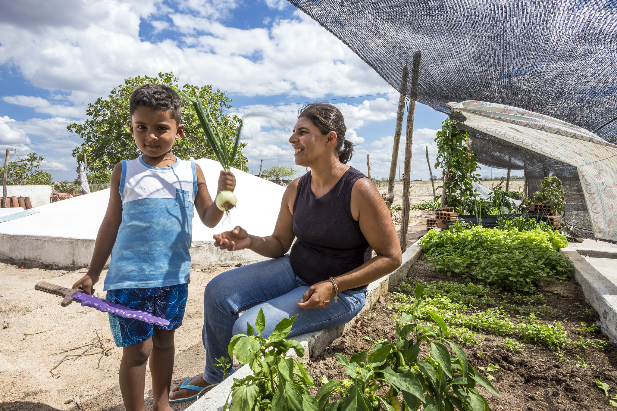 Mulher com criança segura verduras sentada em sua horta, na frente da cisterna calçadão.