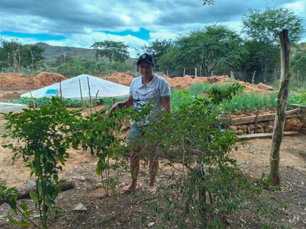 Na foto, a agricultora Maria Nilda pousa ao lado de sua seu quintal produtivo com a cisterna de segunda água atrás.