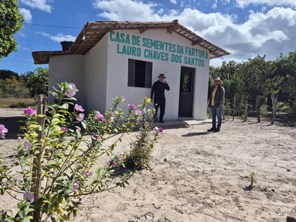 Imagem mostra a fachada branca com letreiro verde escrito Casa de Sementes da Fartura Lauro Chaves dos Santos. Em frente à casa tem dois homens e flores