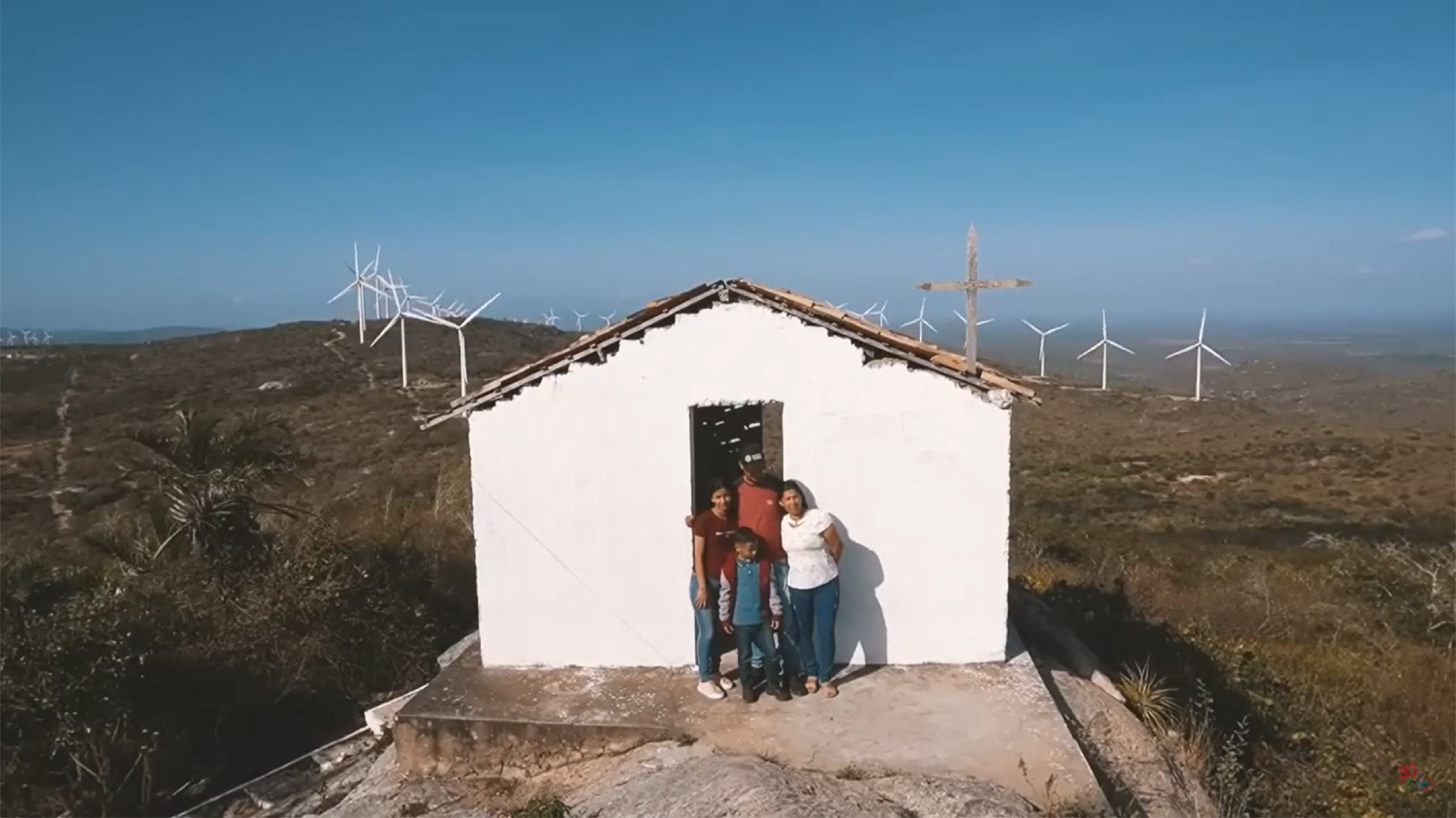 Imagem de uma família composta por um home, uma mulher e uma menina. Eles estão na frente de uma casa branca. Ao fundo, está uma usina de energia eólica, com vários cataventos gigantes. A imagem é do documentário “Vento Agreste”, da Comissão Pastoral da Terra (CPT).