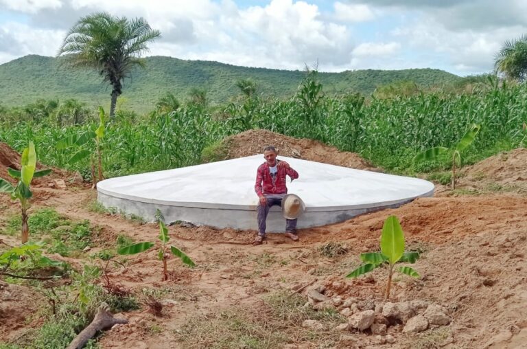 A imagem mostra uma cisterna-calçadão pintada de branco no centro e em cima dela um agricultor sentado