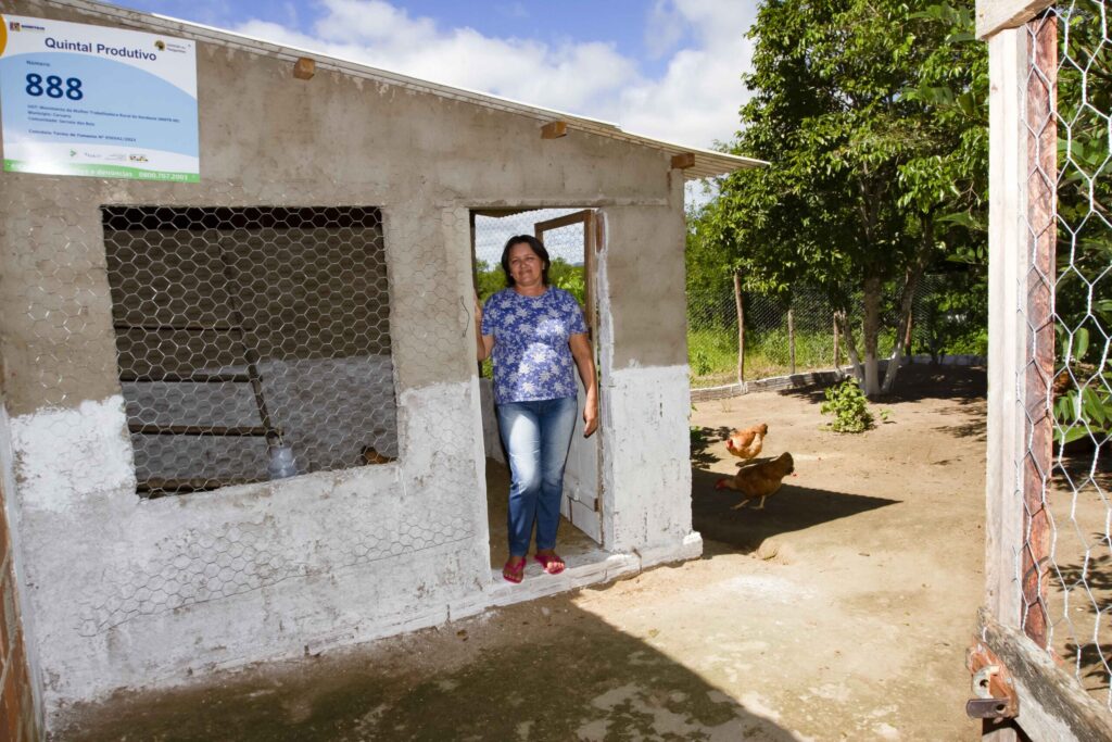 Uma mulher agricultora em frente a uma galinheiro recém construído em alvenaria.