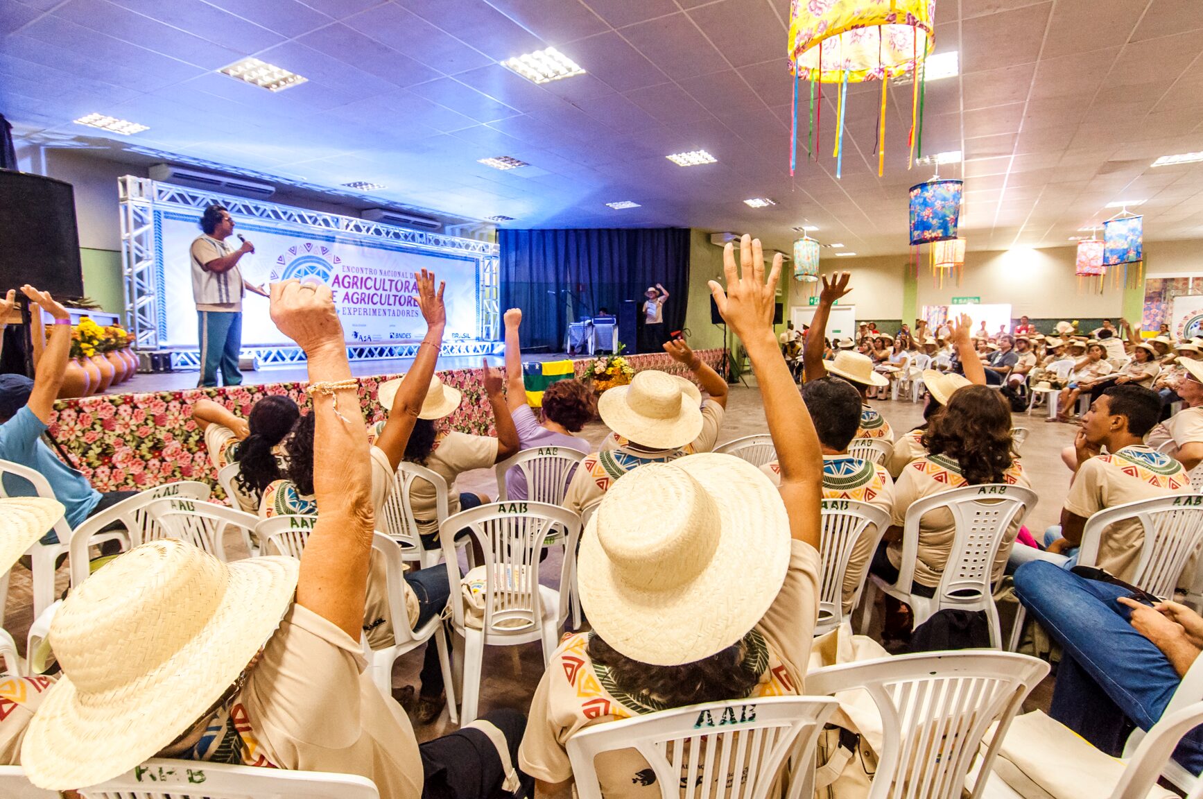 Em uma plenária, vê-se pessoas sentadas com chapéu de palha e mãos levantadas durante o 4º Encontro Nacional de Agricultoras e Agricultores Experimentadores.