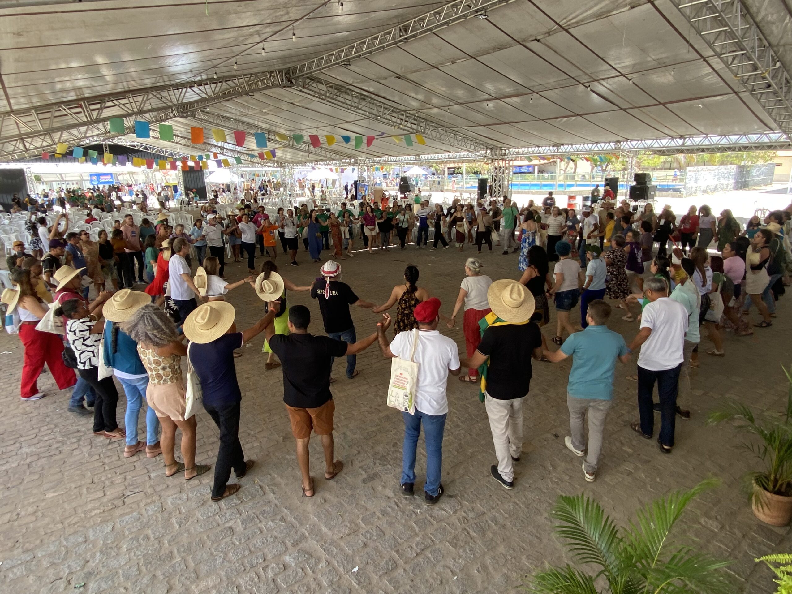 Foto da plenária final do Encontro de agricultores experimentadores. as pessoas participantes estão de mãos dadas em roda dançando uma ciranda.