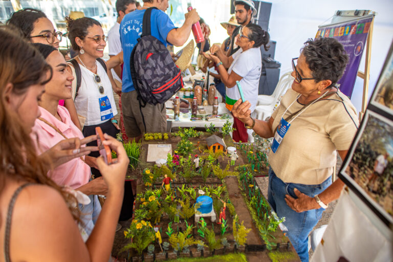imagem de agricultora apresentando a experiência. na frente dela tem uma maquete da área produtiva dela e pessoas conversando.