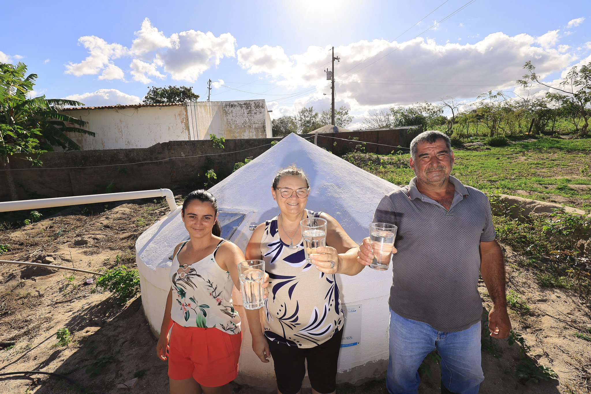 Família de agricultores (pai, mãe filha) seguram copo de água em frente a cisterna, comemorando a tecnologia que lhes garante água para beber e resiliência climática.