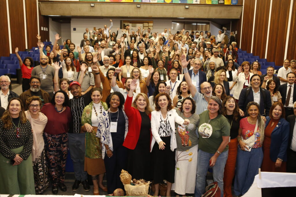 Fotografia em plano aberto mostrando um grande grupo com dezenas de pessoas reunidas em um auditório. Elas estão de pé, posicionadas entre o palco e as fileiras de poltronas azuis ao fundo, que sobem em degraus. A maioria das pessoas sorri e muitas estão com os braços erguidos ou acenando para a câmera, em clima de celebração. Na primeira fila, ao centro.